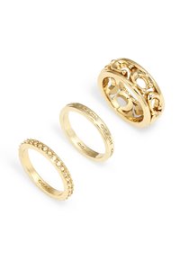 Trois bagues en or avec des designs différents, dont une ornée de cristaux incrustés, une gravée avec "COACH" et une avec des détails découpés.