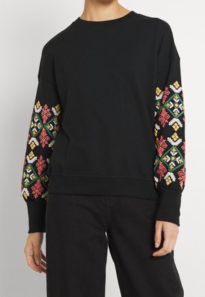 Zwarte top met lange mouwen met meerkleurige geometrische en bloemmotieven borduurwerk op de mouwen, gedragen met een zwarte broek, toont het bovenlichaam en de armen.