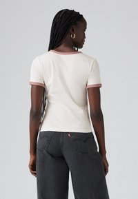 Kvinne med lange fletter iført en hvit t-skjorte med brune detaljer og svarte jeans, som står med ryggen til mot en ensfarget bakgrunn.