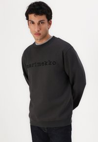 Tmavošedý sveter so zvýšeným logom "marimekko", klasickým okrúhlym výstrihom, dlhými rukávmi a rebrovanými manžetami, štýlový s tmavými džínsy.