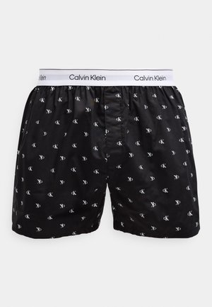 Crne Calvin Klein bokserice s bijelim elastičnim pojasom i malim bijelim logotipima "cK" otisnutim po cijelom proizvodu.