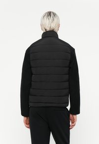 Sort polstret jakke med strukturerede sorte fleeceærmer, der har en stående krave og vandrette sømme. Enkel og funktionel design.