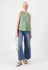 Blusa sem mangas verde clara com decote em V e botões, combinada com calças de ganga azuis de perna larga, sapatos bege e uma mala branca.