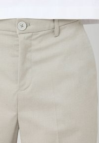 Pantalon beige clair en tissu texturé, doté d'une boutonnière, de poches avant et d'une coupe ajustée avec des détails de couture visibles.