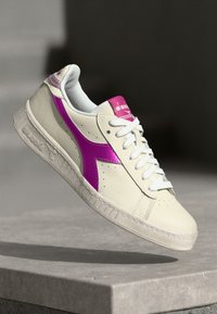 Sneaker with a creamy leather upper, purple accent design, white laces, and textured rubber sole, featuring perforations for breathability.