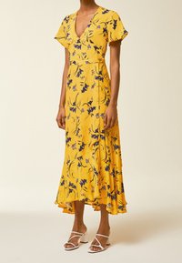 Żółta sukienka midi w kwiaty z falbanistymi rękawami, głębokim dekoltem w kształcie litery V i asymetrycznym dołem. Posiada purpurowe wzory kwiatowe oraz dopasowaną talię.