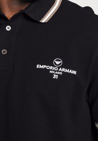Svart polo t-shirt med beige och vit randig krage, vit Emporio Armani Milano 31-logotyp och två knappar på knäppningen.