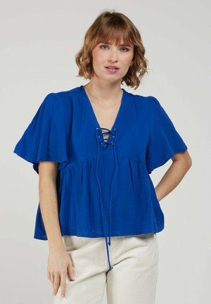 Femme aux cheveux mi-longs portant un chemisier ample bleu roi à col lacé et un pantalon crème, debout devant un fond uni.