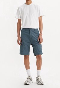 Muž v bílé tričku, modrých cargo šortkách s bočními kapsami, bílých ponožkách do půli lýtek a šedých sportovních teniskách, který stojí na jednobarevném pozadí.