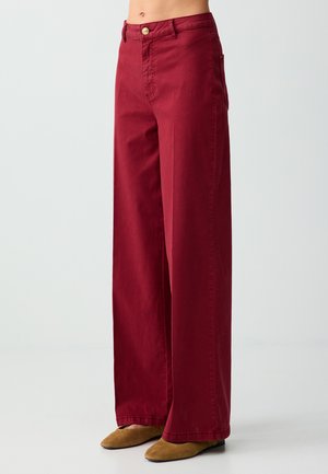 Hoog uitgesneden, wijde bordeauxrode broek met knoop- en ritssluiting, gedragen met lichtbruine puntige platte schoenen op een effen achtergrond.