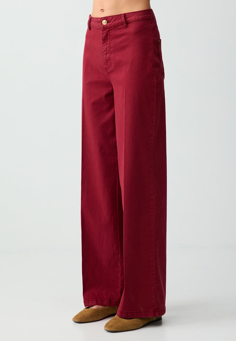 Pantalon bordeaux taille haute à jambes larges avec fermeture par bouton et zip, porté avec des chaussures plates pointues marron clair sur un fond uni.