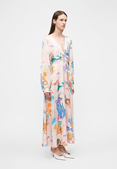 Γυναίκα που στέκεται, φορώντας ένα μακρύ, παστέλ ροζ φόρεμα με πολύχρωμα σχέδια υποβρύχιας θαλάσσιας ζωής και λευκές μυτερές χαμηλές γόβες.