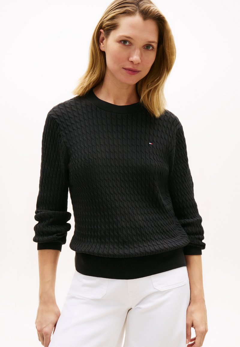 Femme aux cheveux blonds portant un pull noir texturé à manches longues et un pantalon blanc sur un fond clair uni.