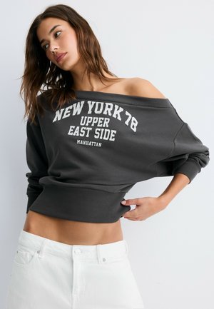 Kvinde med løst brunt hår iført en mørkegrå sweatshirt med bare skuldre med teksten "NEW YORK 78 UPPER EAST SIDE MANHATTAN" og hvide bukser.