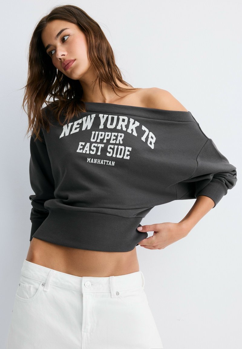 Femme aux cheveux bruns lâches portant un sweat-shirt gris foncé épaules dénudées avec le texte "NEW YORK 78 UPPER EAST SIDE MANHATTAN" et un pantalon blanc.