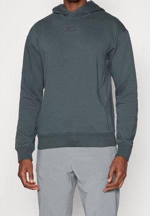 Mann trägt dunkelgrünen Nike Pro Hoodie und hellgraue Nike Cargohose, steht vor einem schlichten weißen Hintergrund.
