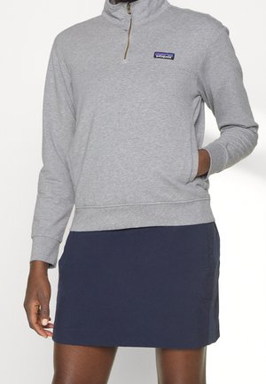 Personne portant un sweat-shirt à fermeture éclair partielle gris clair avec le logo Patagonia et une jupe bleu marine, la main dans la poche du sweat-shirt.