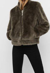 Kvinde iført mørkebrun faux fur zip-up jakke med hænderne i lommerne, parret med sorte bukser.