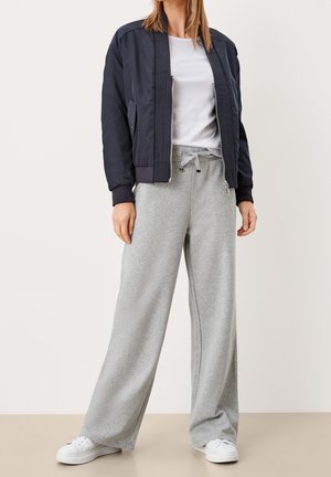 Mujer que lleva una chaqueta bomber azul marino, camiseta blanca, pantalones de chándal grises sueltos con cordón y zapatillas blancas, de pie sobre un suelo beige.