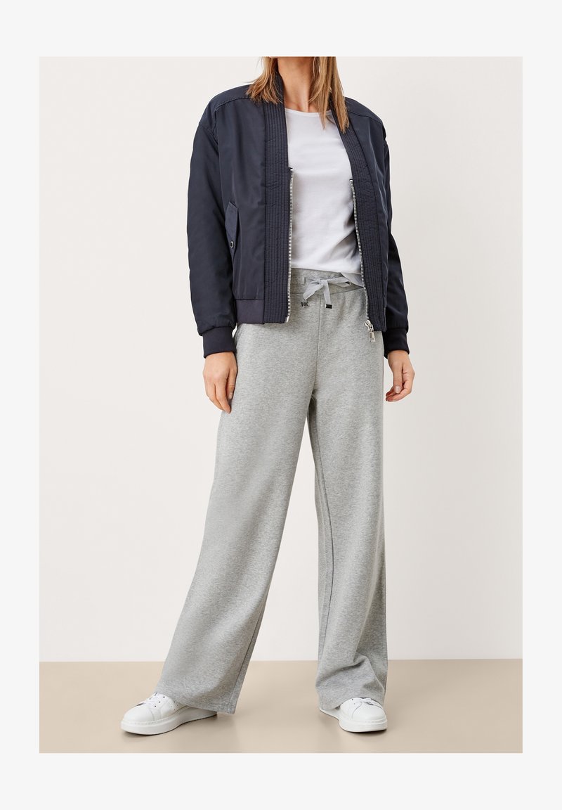 Femme portant une veste bomber bleu marine, un t-shirt blanc, un pantalon de survêtement gris ample avec cordon de serrage et des baskets blanches, debout sur un sol beige.