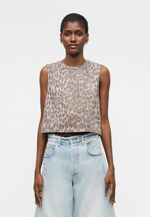 Γυναίκα που φοράει αμάνικη μπλούζα με τύπωμα λεοπάρδαλης και διακοσμημένο ντεκολτέ και ανοιχτό μπλε ψηλόμεσο τζιν με φαρδιά μπατζάκια, στέκεται μπροστά σε απλό φόντο.