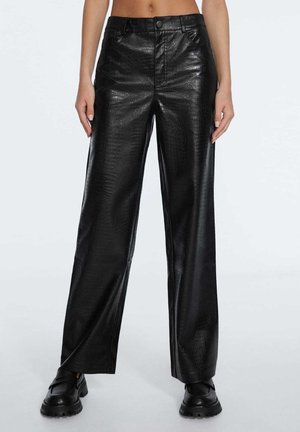 Persoon draagt zwarte high-waisted wijde broek van nepleer met een subtiele krokodillenstructuur en zwarte chunky loafers, rechtopstaand.