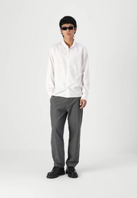Weißes Langarmhemd, graue Hose, schwarze Schuhe und schwarze Sonnenbrille. Das Hemd hat einen Knopfleistenverschluss; die Hose ist leicht locker geschnitten.
