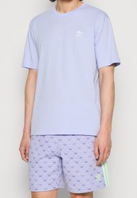 Světle fialové bavlněné tričko s krátkým rukávem a malým bílým logem na hrudi, doplněné vzorovanými šortkami v podobné barevné škále.