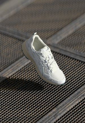Hvid atletisk sko med en tekstureret overdel, snørebåndslukning, gummisål og logonotater, placeret over en tekstureret overflade.