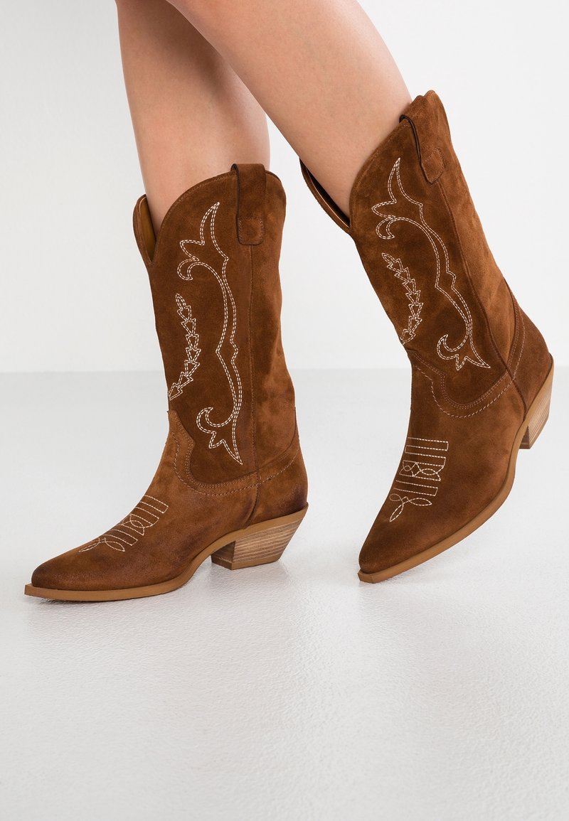 Brune ruskinds cowboy-støvler med hvide syninger, båret på krydsede ben mod en ensfarvet hvid baggrund.