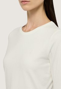 Camisa de manga larga color crema claro hecha de una tela suave. Presenta un cuello redondo y un sutil logo bordado en el pecho. Textura lisa.