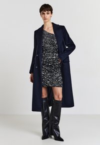 Cappotto lungo blu marino con grandi bottoni sopra un abito corto di paillettes nere e argento. Stivali alti in pelle nera aderenti completano il look.