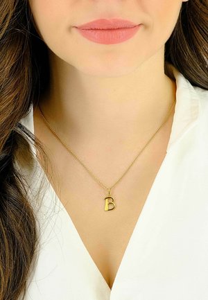 Collana d'oro con ciondolo a forma di "b" minuscola, con una catena sottile, indossata sopra una camicetta bianca. Il design è semplice ed elegante.