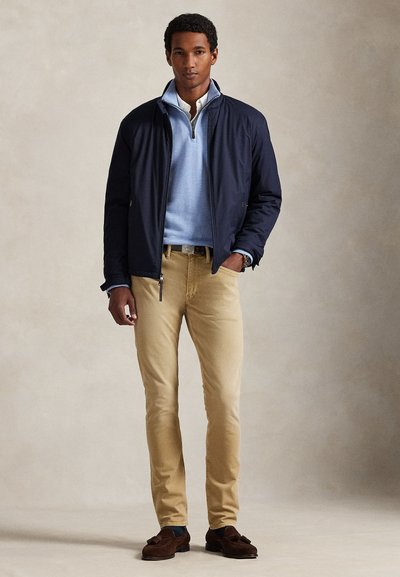 Homme portant une veste bleu marine, un pull léger bleu clair à fermeture éclair jusqu'au quart par-dessus une chemise blanche, un pantalon beige et des mocassins marron, debout avec une main dans la poche.