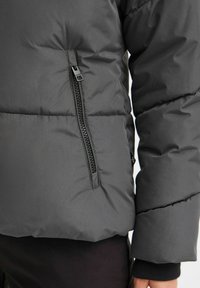 Veste grise matelassée avec une poche zippée et des poignets côtelés. Matériau extérieur lisse avec un surpiqué pour l'isolation.