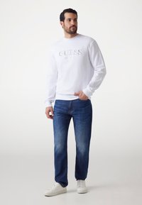 Biała bluza z haftowanym napisem "GUESS EST. 1981", zestawiona z niebieskimi dżinsami i białymi trampkami. Gładki materiał o swobodnym kroju.