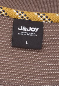 Bruine gestructureerde stof met een geweven gele en marineblauwe ruitrand. Een zwart label bevat witte tekst: "J&JOY", "IEDERE STAP EEN NIEUWE REIS", maat "L".