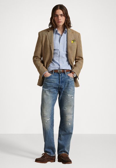 Bruin visnetblazer, lichtblauw overhemd, versleten blauwe jeans, bruine leren riem en donkerbruine schoenen. Een pochette voegt detail toe.
