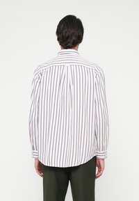 Άνδρας με σκούρα μαλλιά που φοράει ένα λευκό πουκάμισο μακρυμάνικο με κάθετες μπλε και κόκκινες ρίγες, στέκεται γυρισμένος από πίσω, συνδυασμένο με σκούρο πράσινο παντελόνι.