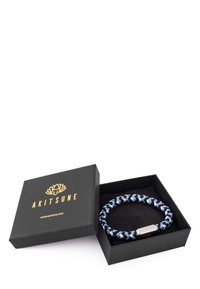 Boîte cadeau noire avec logo Akitsune doré à côté d'un bracelet tressé bleu, noir et blanc avec une fermeture argentée à l'intérieur de la boîte.