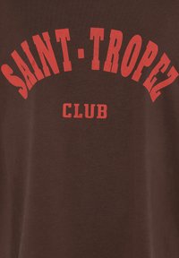 Ruda medžiaga su ryškiomis raudonomis raidėmis, kuriose arkos formoje parašyta "SAINT-TROPEZ" ir žemiau didžiosiomis raidėmis "CLUB".