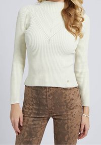 Pull côtelé crème clair à col montant, avec des surpiqûres décoratives en forme de V ; accompagné d'un pantalon marron ajusté à motif.