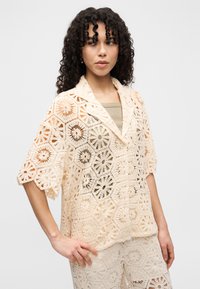 Γυναίκα με σγουρά μαλλιά, φορώντας μπεζ ροχά πλεκτό τοπ και παντελόνι στο ίδιο χρώμα, στέκεται με το χέρι στη μέση, κοιτώντας προς τα πλάγια σε λευκό φόντο.