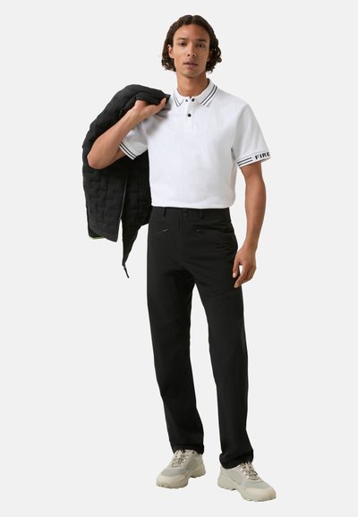 Weißes Poloshirt mit schwarz-weiß gestreiftem Kragen, kombiniert mit schwarzen Hosen und hellgrauen Sneakers. Das Model hält eine schwarze gesteppte Jacke.