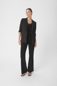 Blazer tailored nero su una maglietta di pizzo nera, abbinato a pantaloni neri svasati e sandali con tacco a strappo. Tessuto liscio, design strutturato.