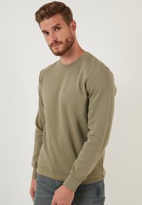 Sudadera de manga larga verde oliva en tejido acanalado con cuello redondo y puños ajustados, combinada con jeans de color azul claro.