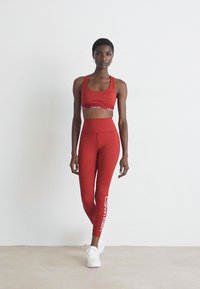 Vrouw in een rode Calvin Klein sportbh en legging met witte sneakers, staand tegen een eenvoudige lichtgekleurde muur en vloer.