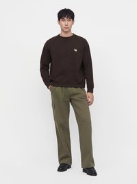 Homme debout portant un sweat-shirt marron foncé avec un petit logo et un pantalon cargo vert olive avec des chaussures noires sur fond blanc uni.