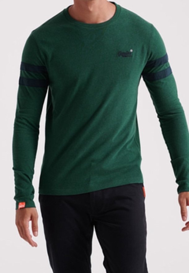 Mann, der ein eng anliegendes grünes Langarmshirt mit schwarzen Streifen an den Ärmeln und schwarzen Hosen trägt, und vor einem schlichten Hintergrund steht.