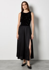 Schwarzes Abendkleid mit einem mit Pailletten verzierten Oberteil, ärmellosem Design und einem fließenden Satinkleid mit einem Seitenschlitz. Flache schwarze Schuhe.